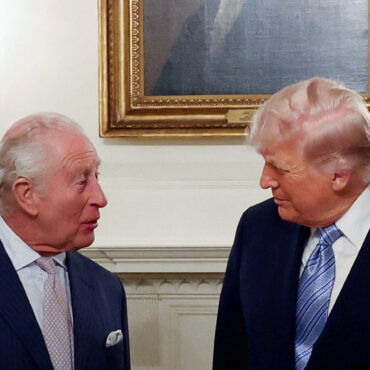 Two older men in dark suits converse in a formal room, facing each other near a fireplace and framed painting.