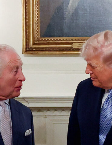 Two older men in dark suits converse in a formal room, facing each other near a fireplace and framed painting.