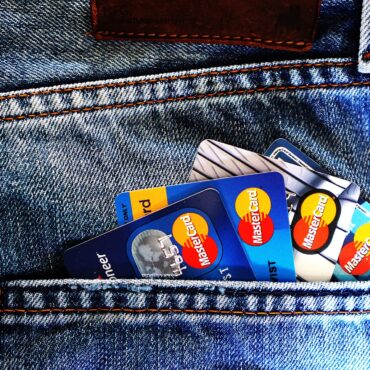 Back pocket of blue denim jeans with several Mastercard credit cards peeking out, orange stitching visible along the pocket edge.