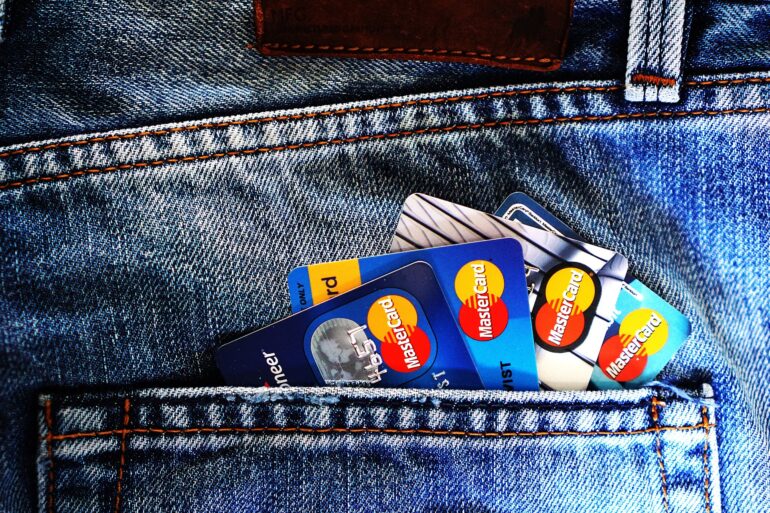 Back pocket of blue denim jeans with several Mastercard credit cards peeking out, orange stitching visible along the pocket edge.