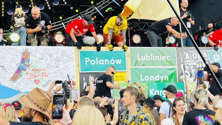 Crowd at an outdoor festival stage, fans reach over the barrier to greet performers while photographers film from above; banners with city names Polanów, Lubliniec, Londyn visible on the barrier.