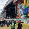 Large outdoor music festival stage with bright graffiti banners and circular lighting rig, crowd gathered in front with flags and signs around the perimeter