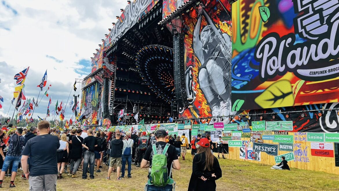 Large outdoor music festival stage with bright graffiti banners and circular lighting rig, crowd gathered in front with flags and signs around the perimeter