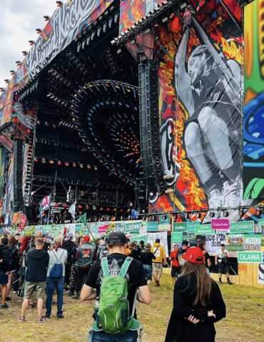 Large outdoor music festival stage with bright graffiti banners and circular lighting rig, crowd gathered in front with flags and signs around the perimeter