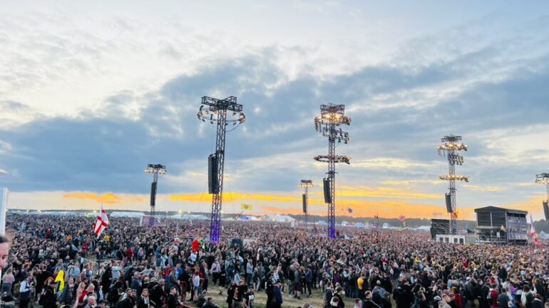 Huge crowd at an outdoor festival at sunset, with tall stage towers and lights rising above the audience.