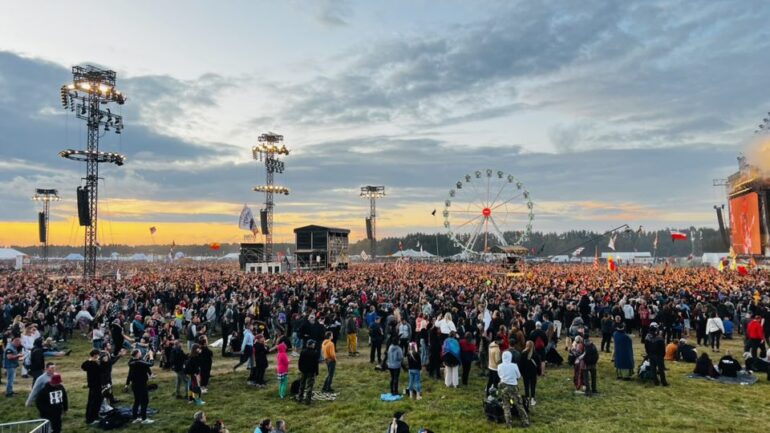 Large crowd at an outdoor music festival at sunset, with tall stage rigs and a distant Ferris wheel.