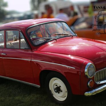 Red vintage car shown from the side and front at a car show, with whitewall tires and chrome trim on a grassy field.