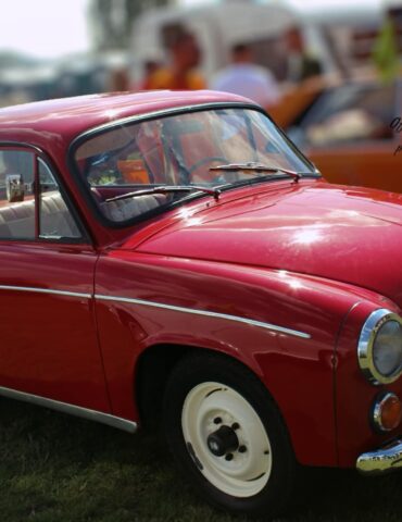 Red vintage car shown from the side and front at a car show, with whitewall tires and chrome trim on a grassy field.