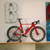 Red road bike on a trainer stand indoors, placed against a wall with a large distressed letter 'B' and a colorful painting to the right.