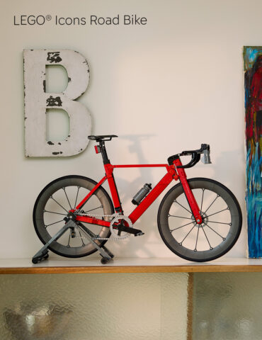 Red road bike on a trainer stand indoors, placed against a wall with a large distressed letter 'B' and a colorful painting to the right.