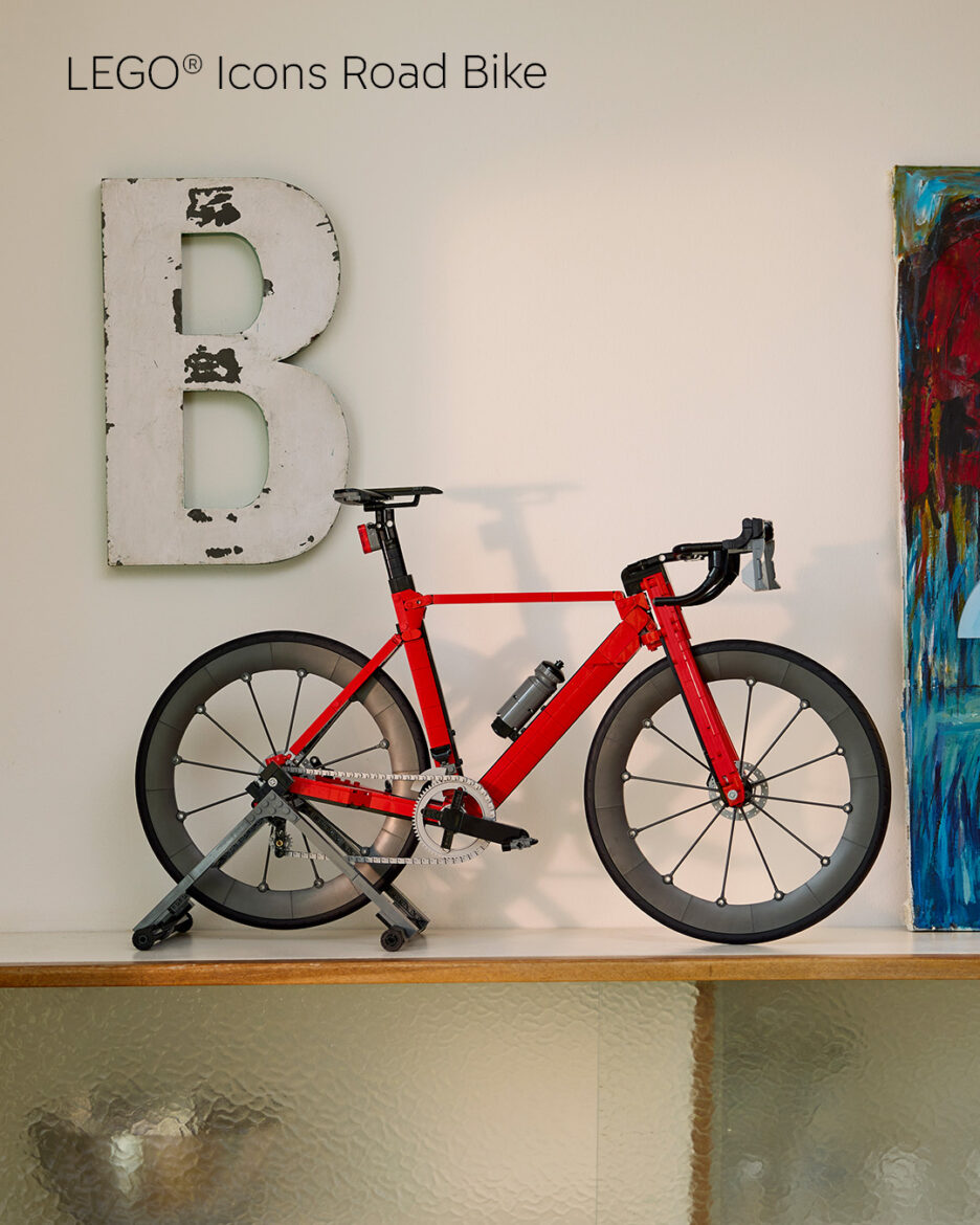 Red road bike on a trainer stand indoors, placed against a wall with a large distressed letter 'B' and a colorful painting to the right.