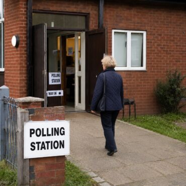Older woman in a navy quilted jacket approaches the open double doors of a brick polling station on a paved path, with a 'Polling Station' sign outside.