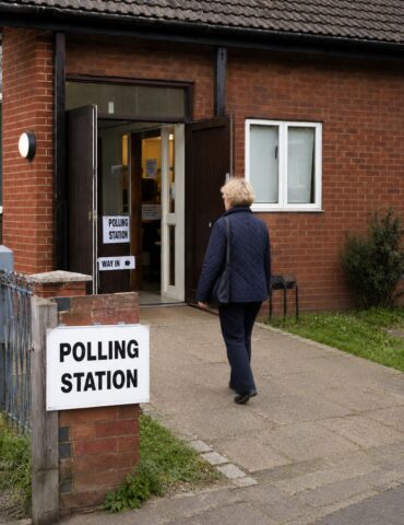 Older woman in a navy quilted jacket approaches the open double doors of a brick polling station on a paved path, with a 'Polling Station' sign outside.