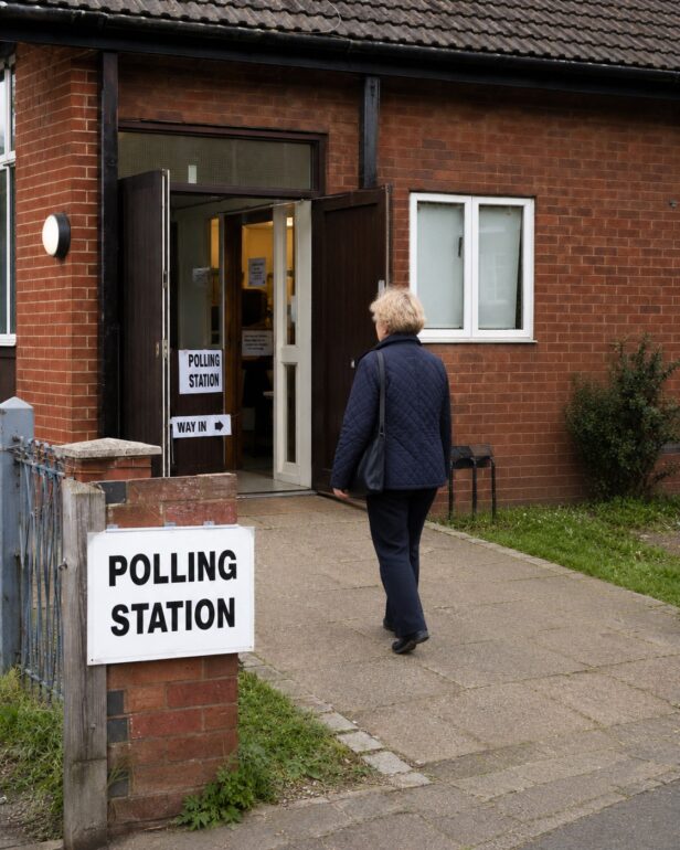 Older woman in a navy quilted jacket approaches the open double doors of a brick polling station on a paved path, with a 'Polling Station' sign outside.