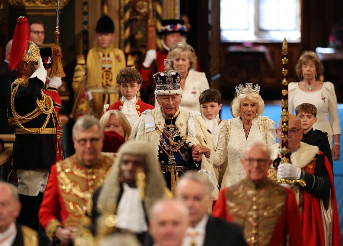 Group of people in ornate royal costumes and crowns gathered indoors for a ceremonial event.