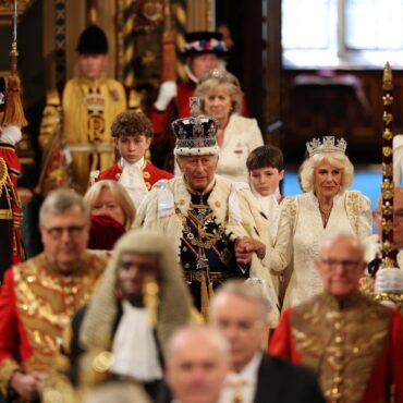 Group of people in ornate royal costumes and crowns gathered indoors for a ceremonial event.