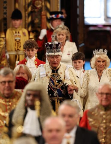 Group of people in ornate royal costumes and crowns gathered indoors for a ceremonial event.