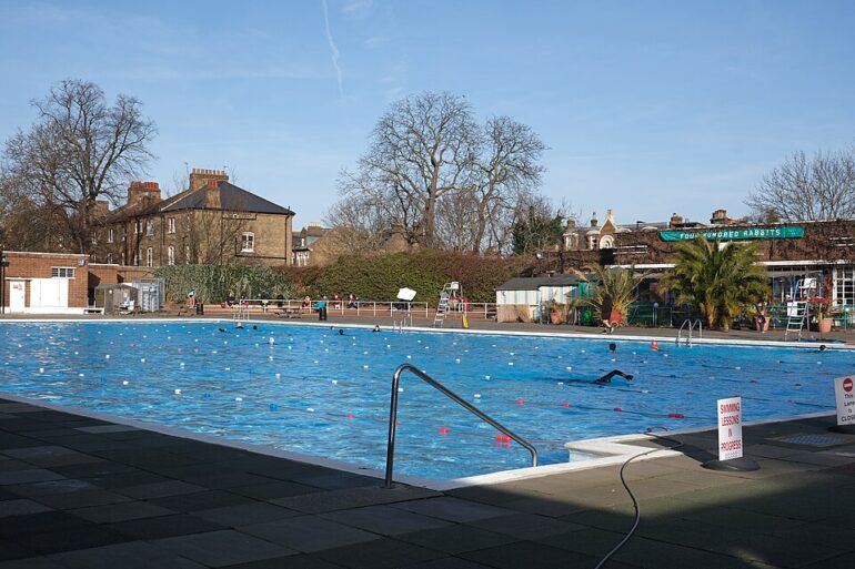 Outdoor public swimming pool on a sunny day; blue water with lane markers, pool deck, and brick buildings in the background at the edge of a residential area.