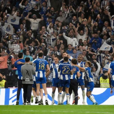 Soccer players in blue-and-white uniforms celebrate a goal on the field with cheering fans in the stands behind them