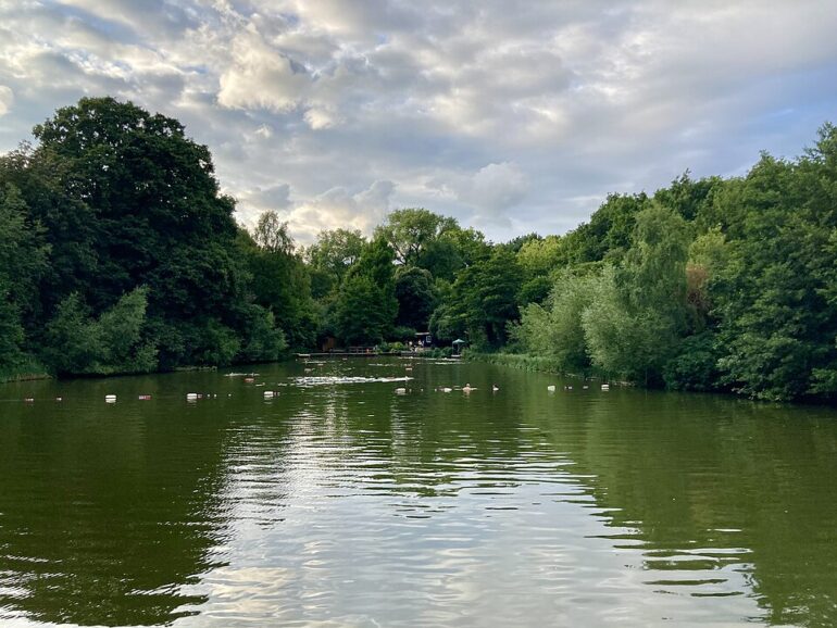 A tranquil lake surrounded by dense green trees, with a cloudy sky and small buoys scattered on the water surface.