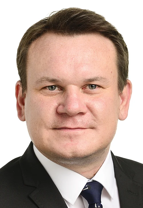 Head-and-shoulders portrait of a smiling man in a dark suit, white shirt, and navy tie.