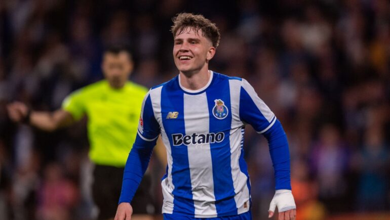 Soccer player in blue-and-white striped kit smiles while running on the field, referee in the background at a crowded stadium.