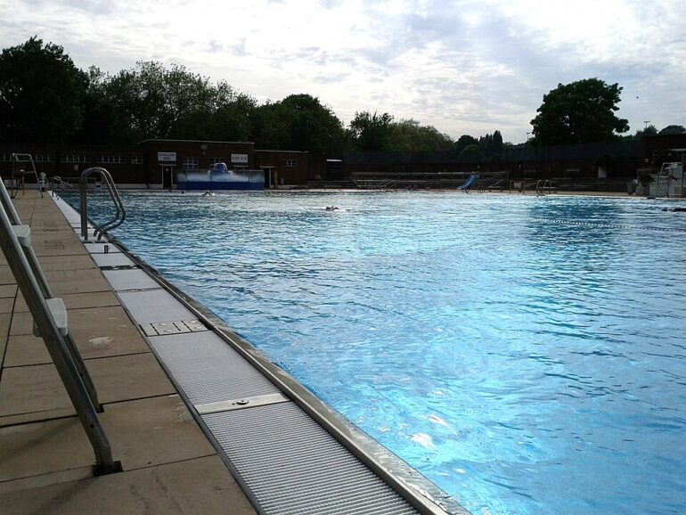 Public outdoor pool with bright blue water, concrete deck, metal ladder on the left, and empty spectator stands in the background.