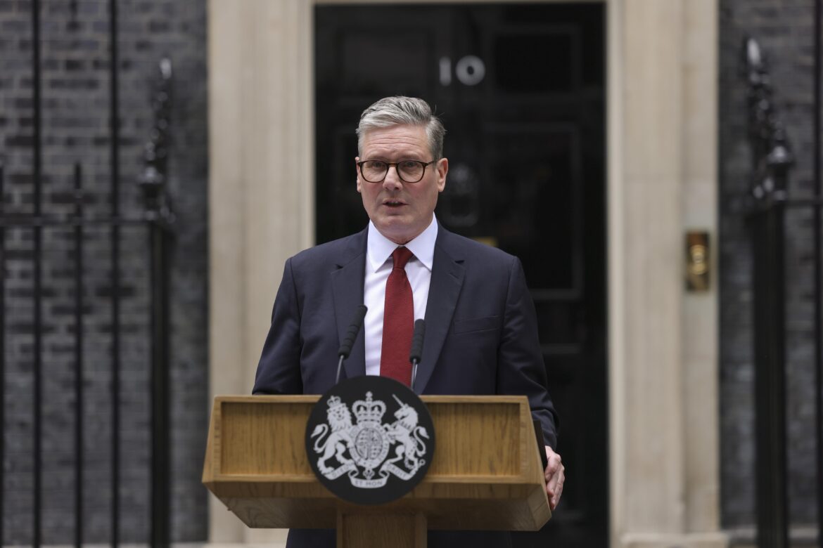 Man in a dark suit and red tie speaks at a wooden podium with a royal crest in front of a government building entrance.