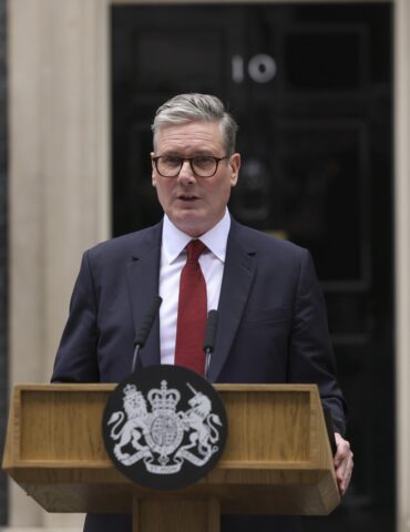 Man in a dark suit and red tie speaks at a wooden podium with a royal crest in front of a government building entrance.