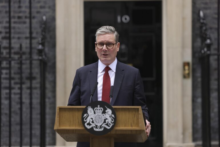 Man in a dark suit and red tie speaks at a wooden podium with a royal crest in front of a government building entrance.