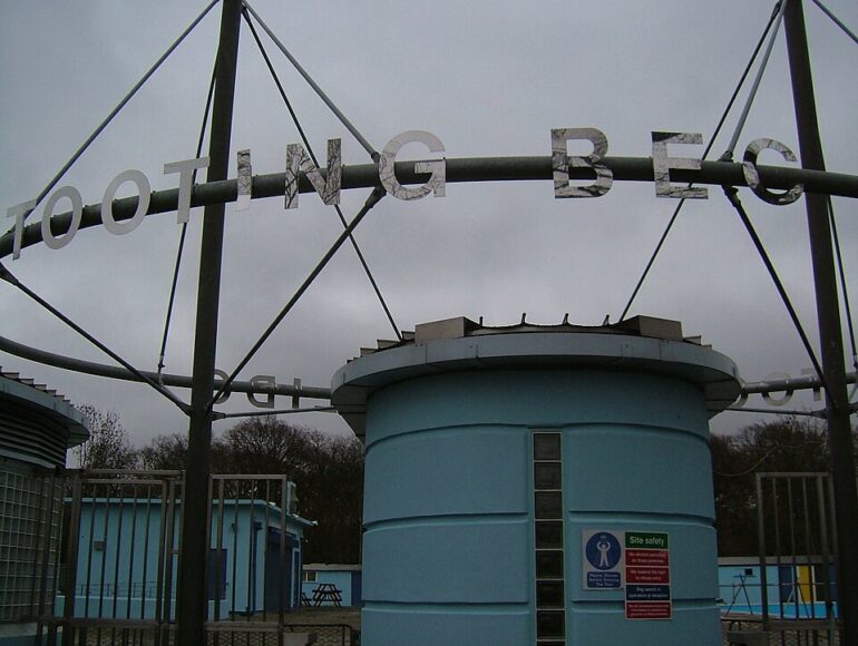 Metal framework arching over a blue cylindrical building, with large letters forming a sign above it; a site-safety sign is mounted on the wall.