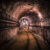Underground tunnel with curved ceiling, rusty rails along a concrete floor, and dim lighting.