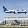 Ryanair Boeing 737 in blue–white livery with yellow harp logo, descending over a terminal building against a clear sky.