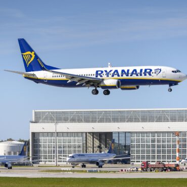 Ryanair Boeing 737 in blue–white livery with yellow harp logo, descending over a terminal building against a clear sky.