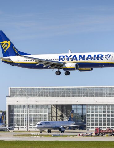 Ryanair Boeing 737 in blue–white livery with yellow harp logo, descending over a terminal building against a clear sky.