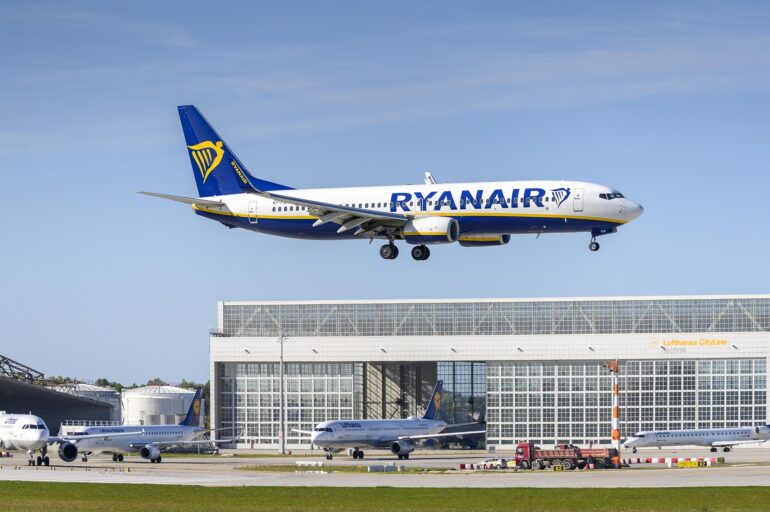 Ryanair Boeing 737 in blue–white livery with yellow harp logo, descending over a terminal building against a clear sky.