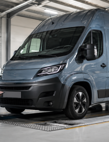 Technician kneeling by a grey van on a workshop lift, inspecting with a tablet in hand.