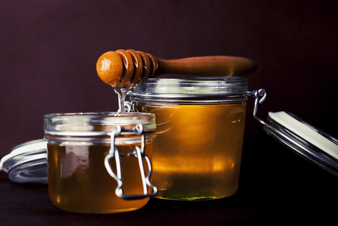 Close-up of glass honey jars with a wooden honey dipper resting on top, honey dripping from the dipper against a dark background