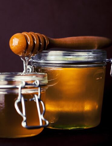 Close-up of glass honey jars with a wooden honey dipper resting on top, honey dripping from the dipper against a dark background