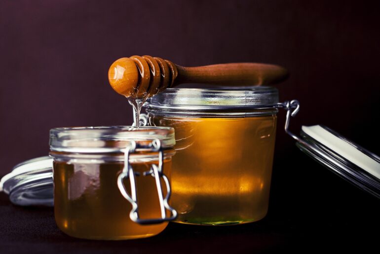 Close-up of glass honey jars with a wooden honey dipper resting on top, honey dripping from the dipper against a dark background