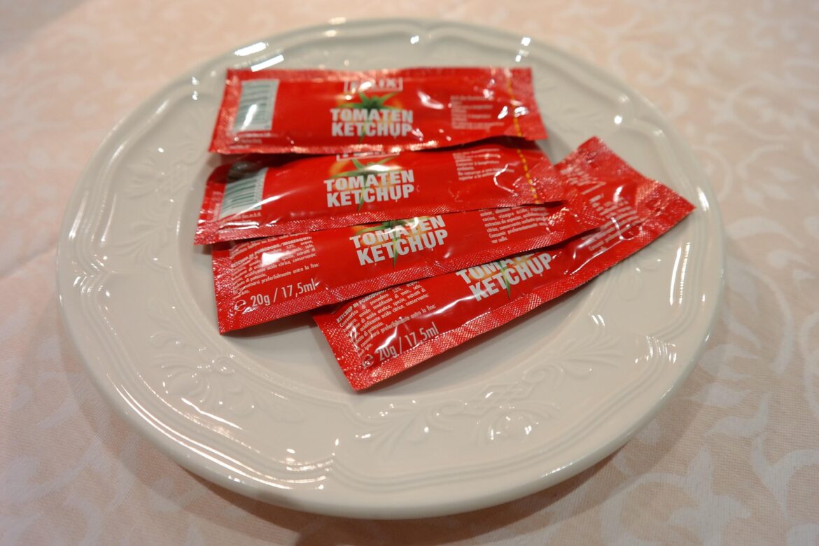 White plate holding several red tomato ketchup packets with text 'Tomaten Ketchup' on a light patterned tablecloth