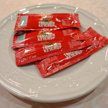 White plate holding several red tomato ketchup packets with text 'Tomaten Ketchup' on a light patterned tablecloth