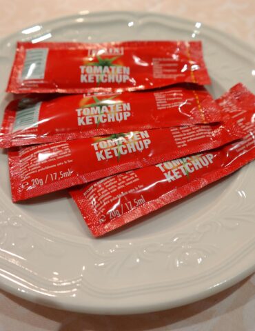 White plate holding several red tomato ketchup packets with text 'Tomaten Ketchup' on a light patterned tablecloth