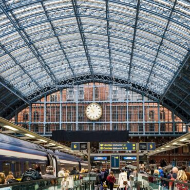 Busy train station platform with a glass-and-steel arched roof, a large central clock, and passengers with luggage.