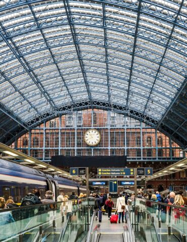 Busy train station platform with a glass-and-steel arched roof, a large central clock, and passengers with luggage.
