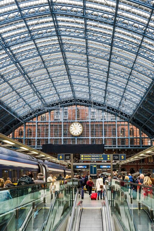 Busy train station platform with a glass-and-steel arched roof, a large central clock, and passengers with luggage.