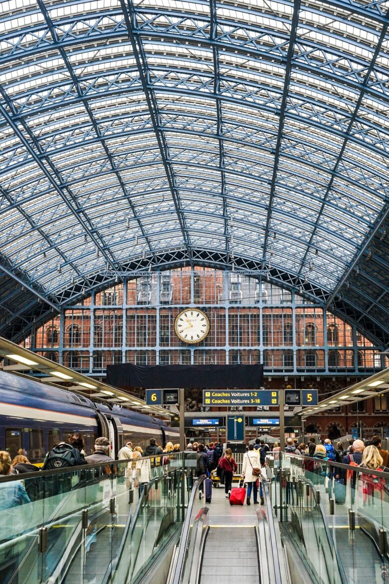 Busy train station platform with a glass-and-steel arched roof, a large central clock, and passengers with luggage.