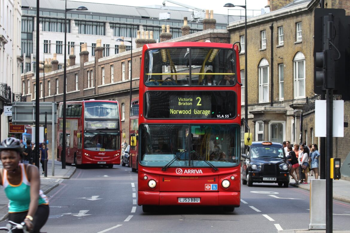 Red London double-decker bus in the foreground on a busy street, route 2 to Victoria Brixton Norwood Garage, with pedestrians nearby.