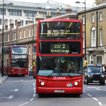 Red London double-decker bus in the foreground on a busy street, route 2 to Victoria Brixton Norwood Garage, with pedestrians nearby.