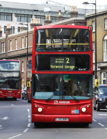 Red London double-decker bus in the foreground on a busy street, route 2 to Victoria Brixton Norwood Garage, with pedestrians nearby.